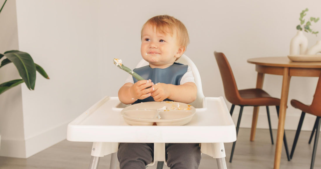 Toddler holding KeaBabies Prep silicone spoon after messy mealtime in high chair.