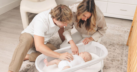 Parents sharing a calm reset moment as their baby sleeps peacefully in a bassinet with a soft fitted bassinet sheet.