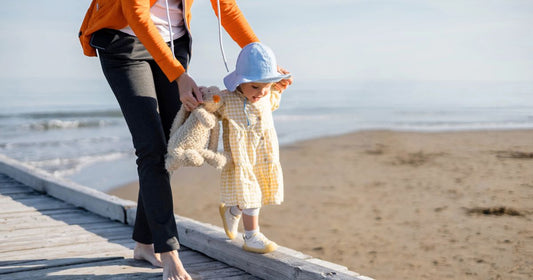 Mom holding a toy while her baby daughter walks on the beach.